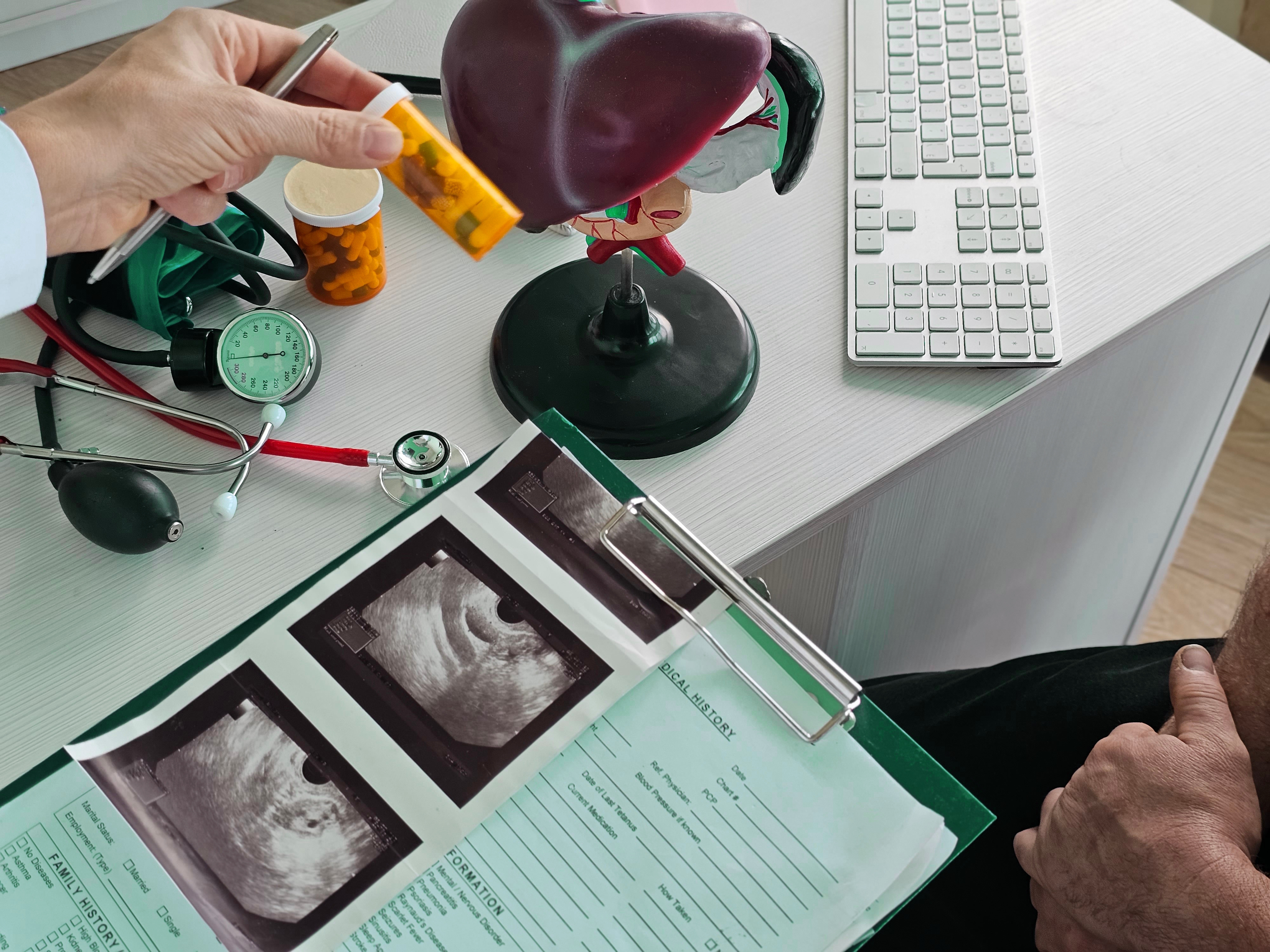 Medical consultation focused on the liver, showing a hand holding a pill bottle, an anatomical liver model, a stethoscope, and printed ultrasound scans on a clipboard